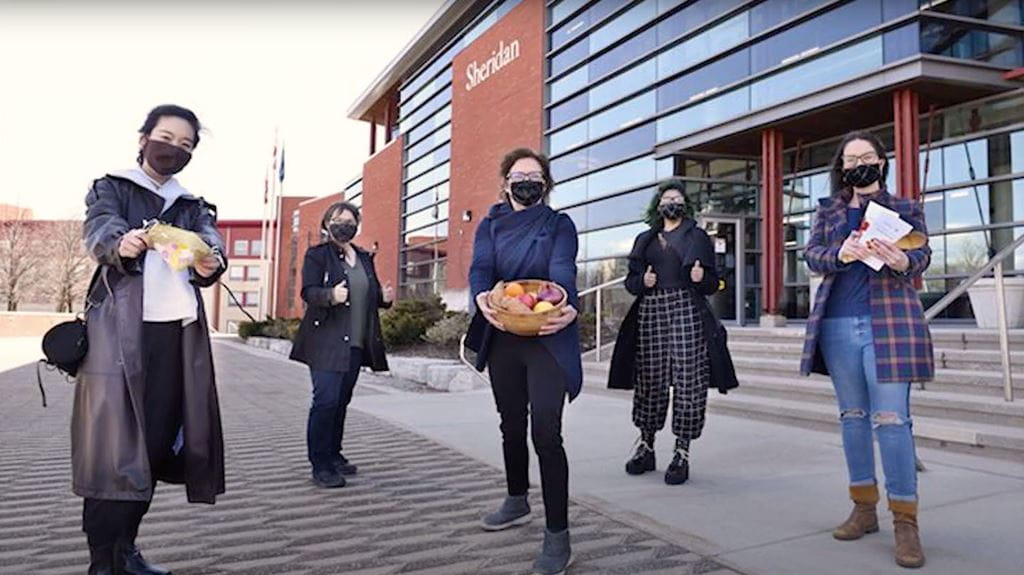 把食物放在桌子上的研究团队戴着口罩，拿着食物站在谢里登特拉法加路校区的一栋建筑前。